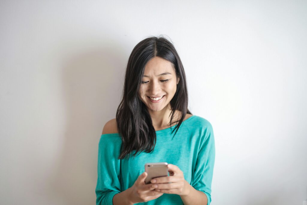 A happy woman in a green top smiling while using her smartphone indoors.